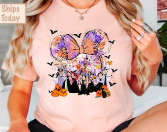 May include: A pink t-shirt with a graphic of a purple and orange bow with a watercolor effect. The bow is surrounded by a group of Disney villains wearing witch hats and holding pumpkins. The text "Ships Today" is in the top left corner of the image.