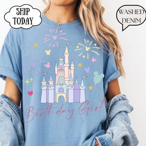 May include: Pale blue t-shirt featuring a castle graphic with fireworks and Mickey Mouse shapes. The words "Birthday Girl" are in cursive. The shirt has a loose fit and the text "Washed Denim" is printed on the shirt.