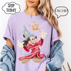 May include: A lilac t-shirt with an embroidered design of Mickey Mouse as a wizard, holding a wand. The design includes a blue wizard hat with stars and a red robe. The text "ORCHID" is in a speech bubble. The words "SHIP TODAY" are in a cloud.