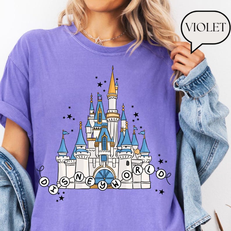May include: A violet-colored t-shirt featuring a whimsical illustration of a castle with blue turrets, gold spires, and the text "DISNEY WORLD" in a circular design. Black stars are scattered around the castle design.