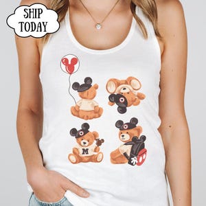 May include: A white tank top featuring a graphic print of four brown teddy bears wearing black mouse ear hats. One bear holds a red balloon, another wears a shirt with the letter M, and one carries a black backpack with red accents. The text SHIP TODAY is visible.