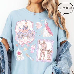 Op de afbeelding: Lichtblauw T-shirt met diverse illustraties van een kasteel, een jurk, ballonnen en een ingelijste afbeelding van een man en een muis. Het shirt heeft het woord "CHAMBRAY" in een tekstballon.