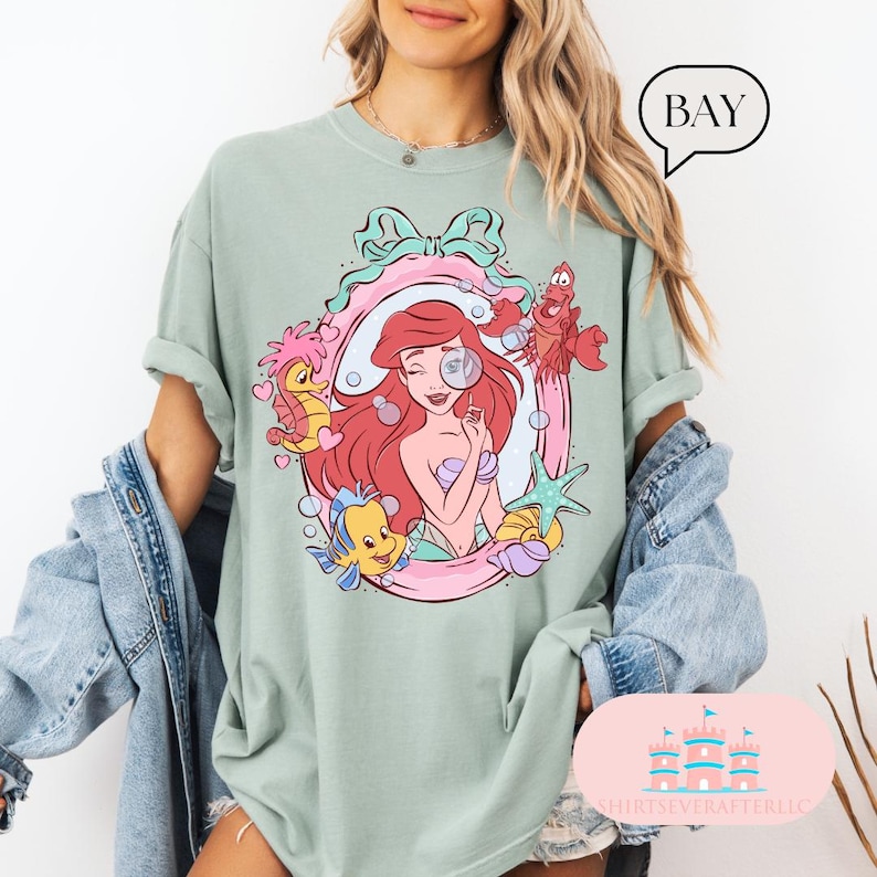 K&ouml;nnte beinhalten: Salbeigr&uuml;nes T-Shirt mit einer farbenfrohen Grafik von Arielle, der Meerjungfrau, umgeben von Meereskreaturen in einem rosa und mintfarbenen Rahmen. Das Wort "BAY" befindet sich in einer Sprechblase. Das T-Shirt hat hochgekrempelte &Auml;rmel.