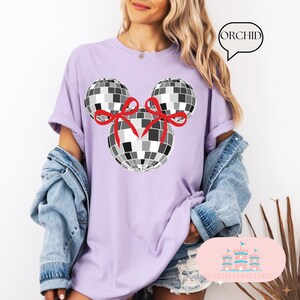 May include: A lavender t-shirt featuring a black and white disco ball Mickey Mouse design. The disco balls have red bows on them. The text "ORCHID" is in a speech bubble.