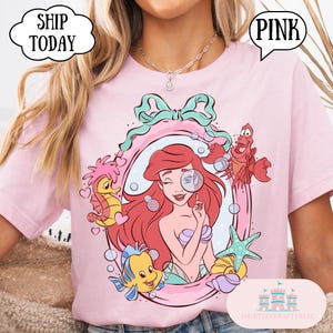 May include: Pink t-shirt featuring a cartoon illustration of Ariel from The Little Mermaid, framed by a teal bow and sea creatures. The word "PINK" is in a speech bubble. The shirt also has the text "SHIP TODAY" in a speech bubble.