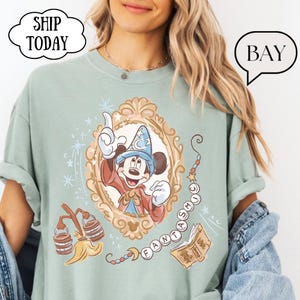 K&ouml;nnte beinhalten: Hellgr&uuml;nes T-Shirt mit einer gerahmten Illustration von Mickey Mouse als Zauberer. Das Design enth&auml;lt das Wort "FANTASTIC" und andere magische Elemente. Das T-Shirt hat hochgekrempelte &Auml;rmel und den Text "SHIP TODAY" in einer wolkenf&ouml;rmigen Blase.