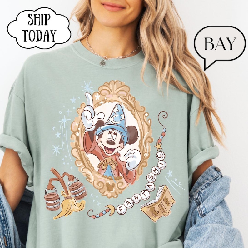 K&ouml;nnte beinhalten: Hellgr&uuml;nes T-Shirt mit einer gerahmten Illustration von Mickey Mouse als Zauberer. Das Design enth&auml;lt das Wort "FANTASTIC" und andere magische Elemente. Das T-Shirt hat hochgekrempelte &Auml;rmel und den Text "SHIP TODAY" in einer wolkenf&ouml;rmigen Blase.