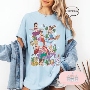 May include: Light blue t-shirt featuring a colorful illustration of mermaids, seaweed, and fish. The graphic includes mermaids with various hair colors and outfits. The word "CHAMBRAY" is in a speech bubble. The shirt is a casual style.
