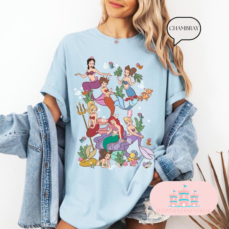 May include: Light blue t-shirt featuring a colorful illustration of mermaids, seaweed, and fish. The graphic includes mermaids with various hair colors and outfits. The word "CHAMBRAY" is in a speech bubble. The shirt is a casual style.