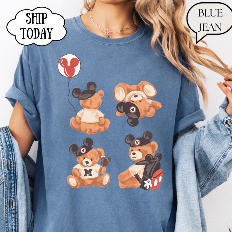 May include: A blue jean colored t-shirt featuring a graphic print of four brown teddy bears wearing black mouse ear hats. One bear holds a red balloon, one is upside down, one wears a shirt with the letter M, and one carries a backpack.