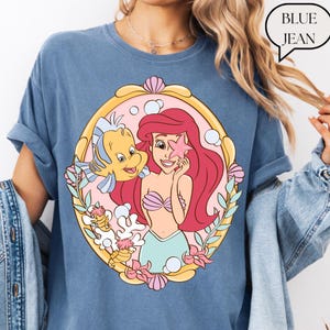 May include: Blue jean colored t-shirt featuring a cartoon illustration of a mermaid holding a starfish, with a yellow fish. The illustration is framed in a gold oval with pink and coral accents. The text "BLUE JEAN" is in a speech bubble.