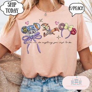May include: Peach-colored t-shirt featuring cartoon characters and accessories. The design includes a police badge, a carrot, a fox, and a bow. The text "You can be anything you wish to be" is also present.