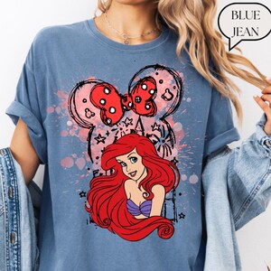 May include: Blue jean colored t-shirt featuring a graphic of Ariel from The Little Mermaid with a red bow and Mickey Mouse ears. The text "BLUE JEAN" is in a speech bubble.