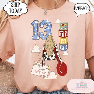 May include: A peach-colored t-shirt featuring a graphic of a person with long blonde hair, a cow-print jacket with red fringe, and a red hat. The design includes a blue cloud-patterned number 13 balloon, alphabet blocks, and a sign reading TAY'S ROOM KEEP OUT except you travis.