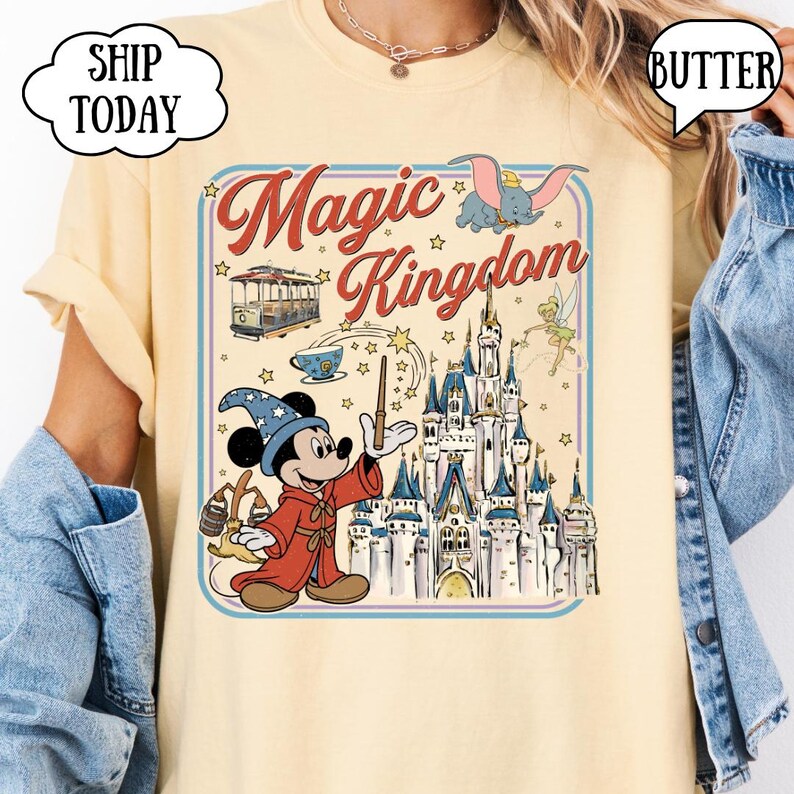 May include: A light yellow t-shirt featuring a colorful graphic of Mickey Mouse as a sorcerer, the Magic Kingdom logo, and other Disney characters. The design includes a castle, Dumbo, and Tinkerbell.