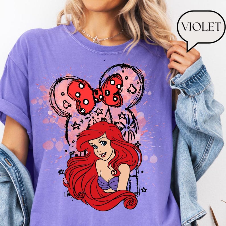 May include: A violet t-shirt featuring a graphic of Ariel from The Little Mermaid, with a red bow and Mickey Mouse ears design. The shirt has a relaxed fit and short sleeves. The word "VIOLET" is in a speech bubble.