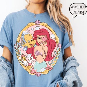 May include: Light blue t-shirt featuring a colorful graphic of Ariel from The Little Mermaid, with Flounder. The design is framed in a gold oval with pink and yellow accents. The text "Washed Denim" is in a speech bubble.