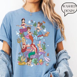 Op de afbeelding: Lichtblauw T-shirt met een kleurrijke illustratie van zeemeerminnen en zeedieren. Het ontwerp toont verschillende zeemeerminnen met verschillende haar- en staartkleuren, omringd door waterplanten en vissen. De tekst "Washed Denim" staat in een tekstballon.