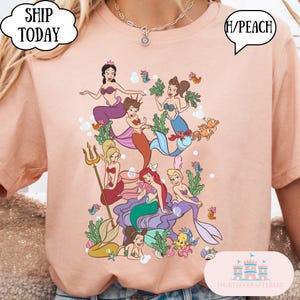 Op de afbeelding: Perzikkleurig T-shirt met een kleurrijke illustratie van verschillende zeemeerminnen met verschillende haarkleuren en outfits. Het ontwerp bevat een drietand, vissen en zeewier. Tekst op het shirt zegt "SHIP TODAY" en "H/PEACH".