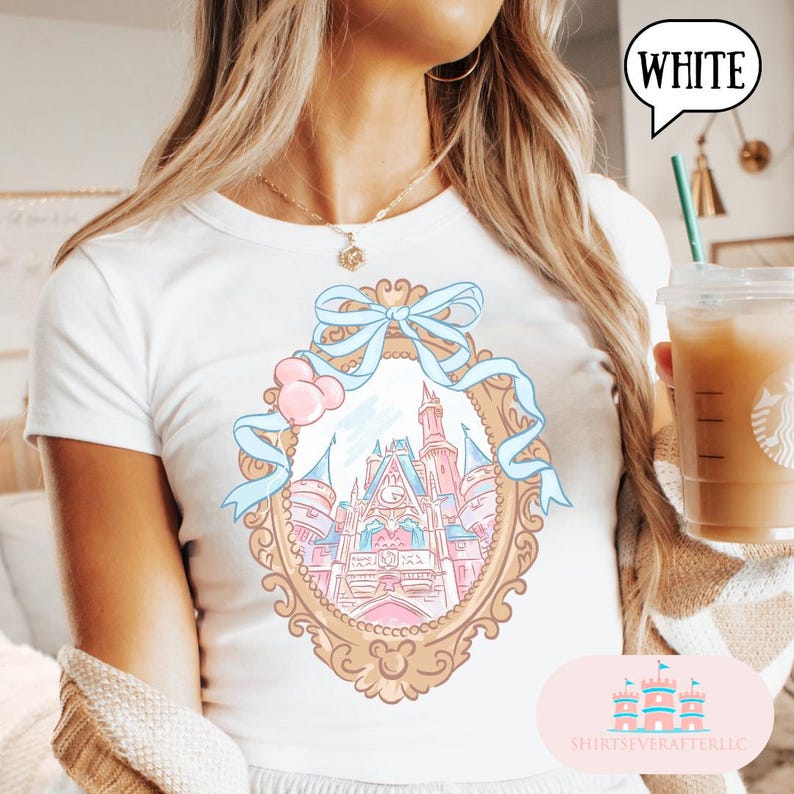 Puede incluir: Camiseta blanca con un dise&ntilde;o de castillo en colores pastel dentro de un marco dorado, adornada con una cinta azul y globos rosas. La camiseta tiene cuello redondo y mangas cortas. La palabra "WHITE" est&aacute; en una burbuja.
