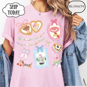 Op de afbeelding: Roze T-shirt met kleurrijke cartoonafbeeldingen van personages en elementen uit een populaire animatiefilm. Het shirt heeft ingelijste afbeeldingen, strikken en de zin "To Infinity and Beyond". De tekst "Ship Today" en "Blossom" zijn ook zichtbaar.