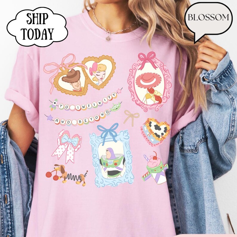 Op de afbeelding: Roze T-shirt met kleurrijke cartoonafbeeldingen van personages en elementen uit een populaire animatiefilm. Het shirt heeft ingelijste afbeeldingen, strikken en de zin "To Infinity and Beyond". De tekst "Ship Today" en "Blossom" zijn ook zichtbaar.