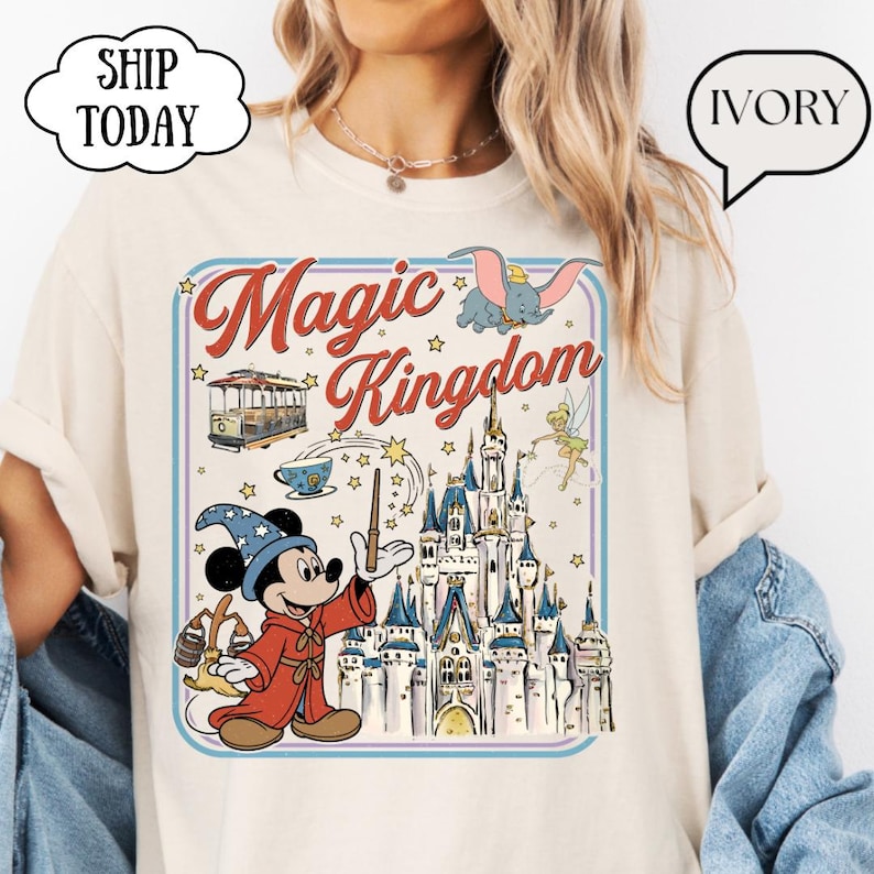 May include: Ivory-colored t-shirt featuring a colorful graphic of Mickey Mouse in a wizard outfit, a castle, and the words "Magic Kingdom." The design includes Dumbo, Tinkerbell, and a trolley car. The shirt has a crew neck and short sleeves.