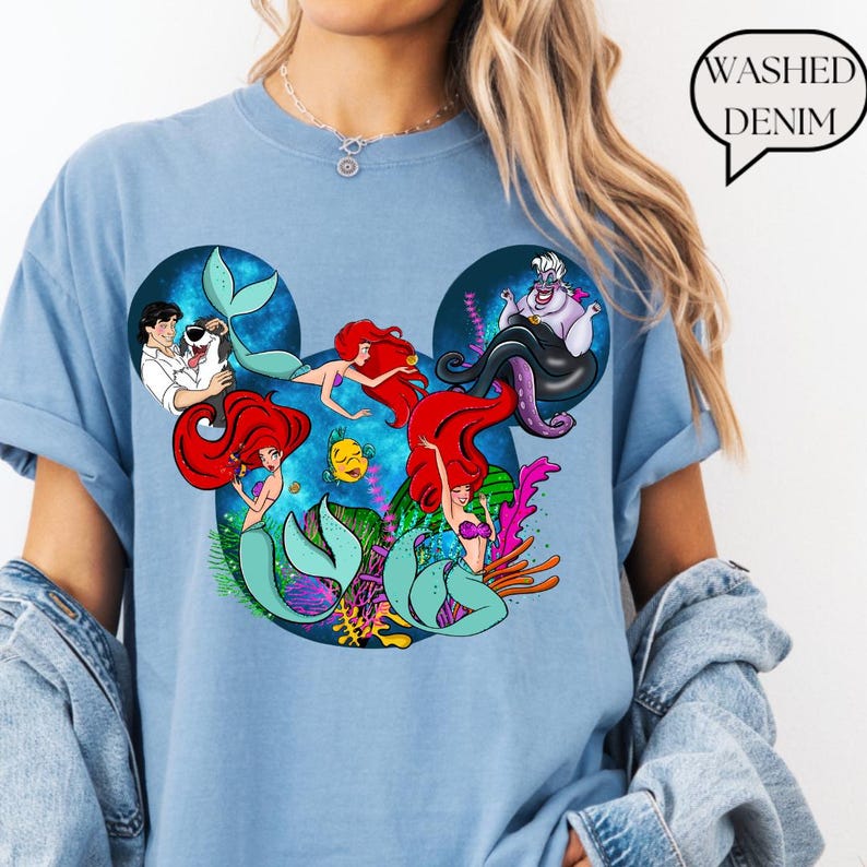 Puede incluir: Camiseta azul claro con un gr&aacute;fico de los personajes de La Sirenita de Disney. El dise&ntilde;o incluye a Ariel, &Uacute;rsula y otros personajes con forma de orejas de Mickey Mouse. El texto "Washed Denim" est&aacute; en una burbuja.