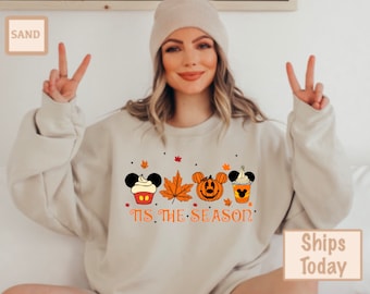 May include: Beige sweatshirt with a fall-themed graphic. The design includes a cupcake, a maple leaf, a jack-o'-lantern, and a coffee drink, all with Mickey Mouse ears. The text "Tis the Season" is printed below the graphics.
