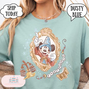 K&ouml;nnte beinhalten: Staubblaues T-Shirt mit einer gerahmten Illustration von Mickey Mouse in einem Zaubererhut, mit dem Wort "FANTASMIC" und anderen magischen Elementen. Das Shirt hat auch den Text "SHIP TODAY" und "DUSTY BLUE".