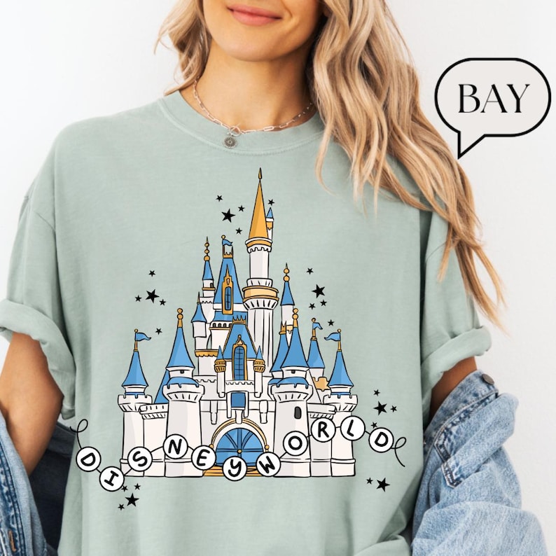 May include: A light green t-shirt featuring a detailed illustration of a fairytale castle with blue turrets and gold accents. The text "DISNEY WORLD" is written below the castle in a circular design, with black stars scattered around the design.