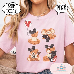 May include: A light pink t-shirt featuring a graphic print of four brown teddy bears wearing black mouse ear hats. One bear holds a red balloon, another is upside down, one wears a shirt with an M, and one carries a black backpack with red accents.