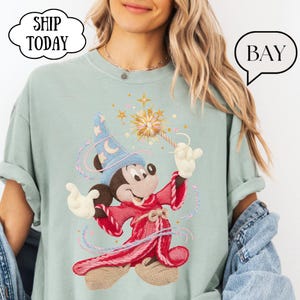 May include: A light green t-shirt featuring an embroidered Mickey Mouse dressed as a wizard, holding a wand with a starburst design. The shirt has the text "SHIP TODAY" in a cloud-shaped bubble.