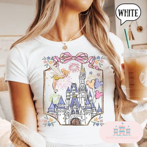 Puede incluir: Camiseta blanca con un gr&aacute;fico colorido de un castillo de cuento de hadas con fuegos artificiales, Campanilla y globos de Mickey Mouse. El dise&ntilde;o est&aacute; enmarcado por un lazo rosa y detalles florales. La palabra "WHITE" est&aacute; en una burbuja.