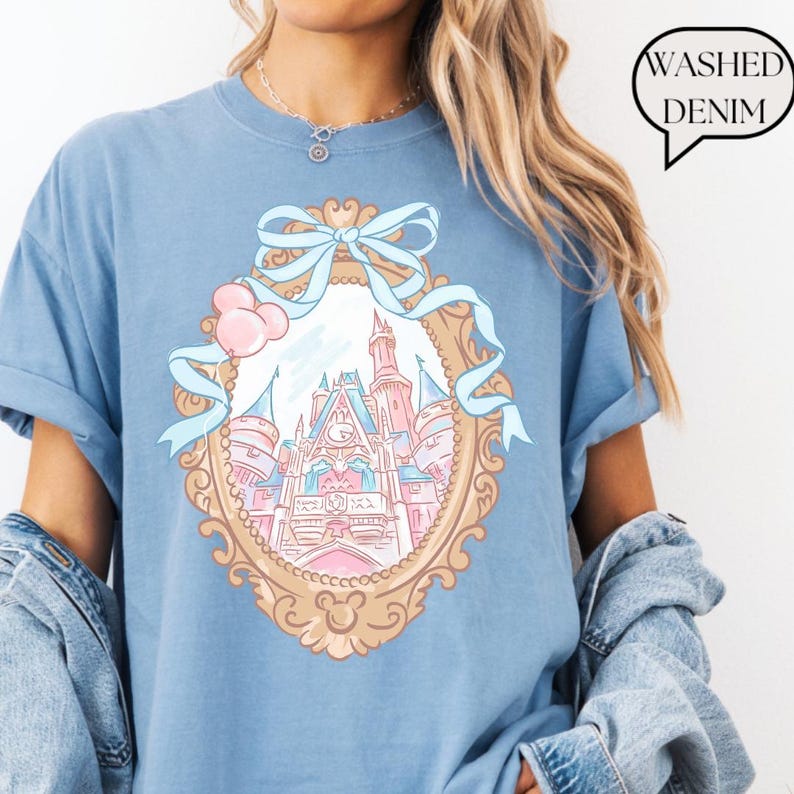 Puede incluir: Camiseta azul claro con una ilustraci&oacute;n de un castillo de cuento de hadas dentro de un marco decorativo, adornada con un lazo azul claro y detalles rosas. El texto "Washed Denim" est&aacute; en un bocadillo.