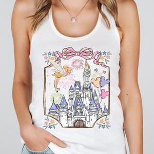 Puede incluir: Camiseta sin mangas blanca con una colorida ilustraci&oacute;n de un castillo de cuento de hadas, fuegos artificiales y Campanilla. El dise&ntilde;o incluye un lazo rosa, globos de Mickey Mouse y detalles florales. Una camiseta gr&aacute;fica caprichosa.