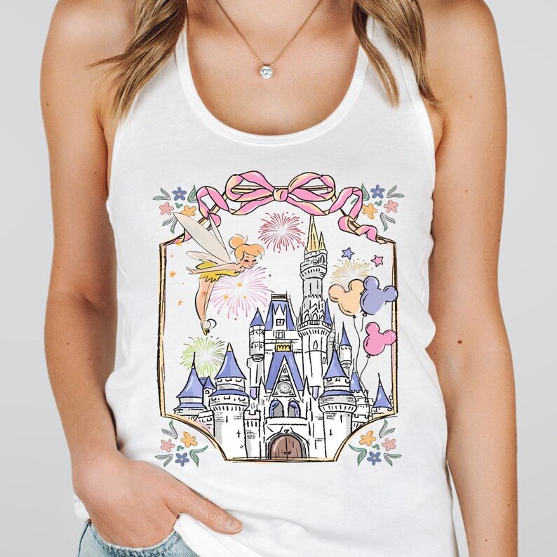 Puede incluir: Camiseta sin mangas blanca con una colorida ilustraci&oacute;n de un castillo de cuento de hadas, fuegos artificiales y Campanilla. El dise&ntilde;o incluye un lazo rosa, globos de Mickey Mouse y detalles florales. Una camiseta gr&aacute;fica caprichosa.