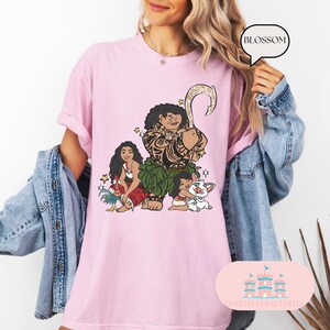 May include: Pink t-shirt with a graphic of Maui, Moana, and Pua from the Disney movie Moana. The graphic is in a cartoon style and features Maui with his fish hook, Moana with her hair in a braid, and Pua the pig.