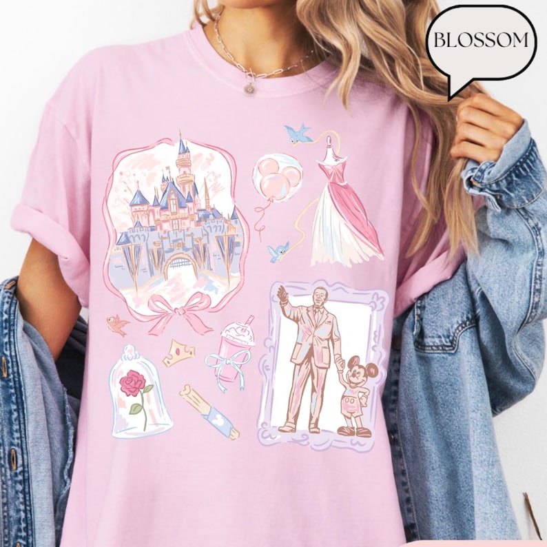 Op de afbeelding: Lichtroze T-shirt met diverse illustraties, waaronder een kasteel, een jurk en een man met Mickey Mouse. Het shirt heeft het woord "BLOSSOM" in een tekstballon.