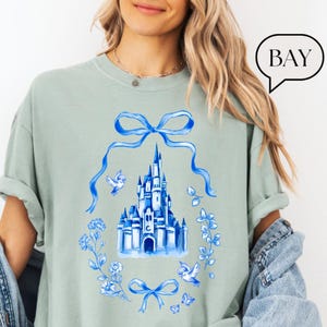 以下が含まれることがあります： 水色のTシャツ。青い水彩画の城のデザインで、青いリボン、鳥、花の要素に囲まれています。「BAY」という言葉が右上にある吹き出しの中にあります。