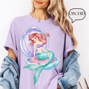 May include: A lilac t-shirt with a cartoon mermaid design. The mermaid, with red hair and a teal tail, is seated in a seashell. The word "ORCHID" is in a speech bubble. The shirt has rolled-up sleeves.