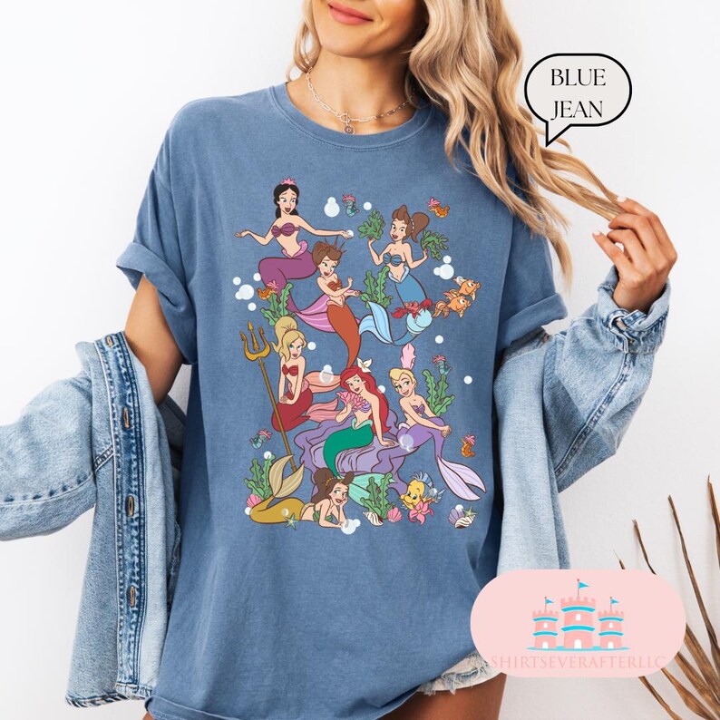May include: Blue jean colored t-shirt featuring a colorful illustration of mermaids. The design includes various mermaids with different hair colors and mermaid tails, surrounded by aquatic elements. The text "BLUE JEAN" is in a speech bubble.