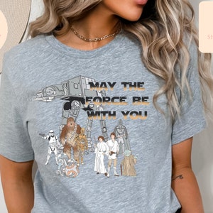 Puede incluir: Camiseta gris con un gr&aacute;fico de personajes de Star Wars, incluyendo Chewbacca, Darth Vader, Yoda y R2-D2. El texto "Que la fuerza te acompa&ntilde;e" est&aacute; impreso en la camiseta.