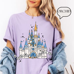 May include: A light purple t-shirt featuring a detailed illustration of a fairytale castle with blue turrets and gold accents. The text "DISNEY WORLD" is arched below the castle. Black stars are scattered around the castle.