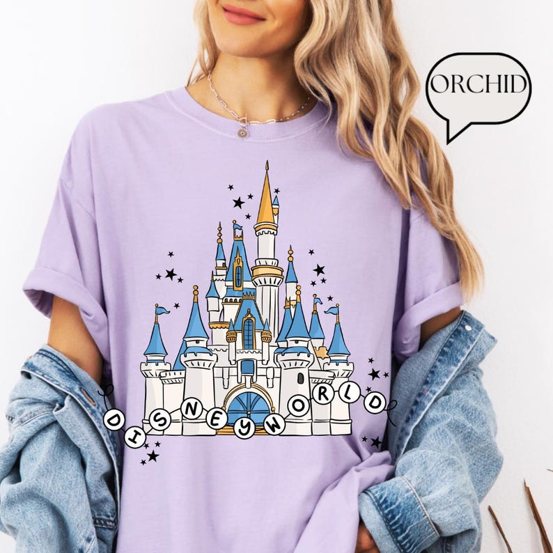 May include: A light purple t-shirt featuring a detailed illustration of a fairytale castle with blue turrets and gold accents. The text "DISNEY WORLD" is arched below the castle. Black stars are scattered around the castle.