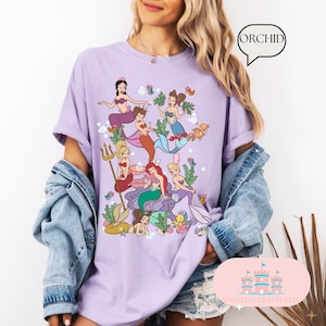 May include: A lilac-colored t-shirt featuring a colorful illustration of mermaids. The graphic includes various mermaids with different hair colors and mermaid tails, surrounded by aquatic plants and sea creatures. The word "ORCHID" is in a speech bubble.