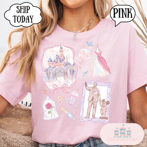 Op de afbeelding: Roze T-shirt met Disney-thema illustraties, waaronder een kasteel, jurk en personages. Het shirt heeft het woord "PINK" in een tekstballon en de woorden "SHIP TODAY" in een wolk-vormige ballon.
