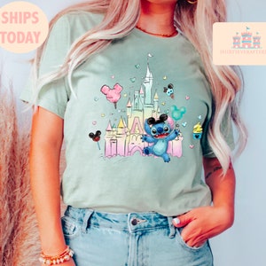 May include: A light green t-shirt with a pastel-colored illustration of a cartoon Stitch character holding a balloon in front of a pastel-colored castle. The castle is surrounded by hearts and other cartoon details.