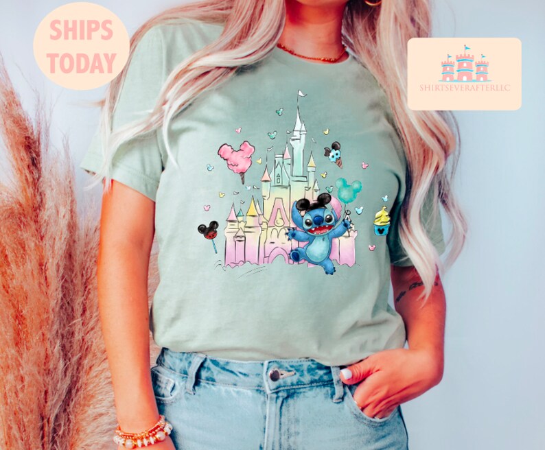 May include: A light green t-shirt with a pastel-colored illustration of a cartoon Stitch character holding a balloon in front of a pastel-colored castle. The castle is surrounded by hearts and other cartoon details.