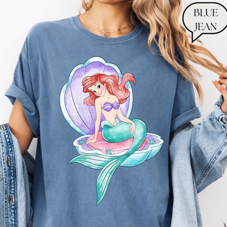 May include: A blue jean coloured t-shirt with a colourful illustration of a mermaid in a seashell. The mermaid has red hair, a purple seashell bra, and a teal tail. The text "BLUE JEAN" is in a speech bubble.